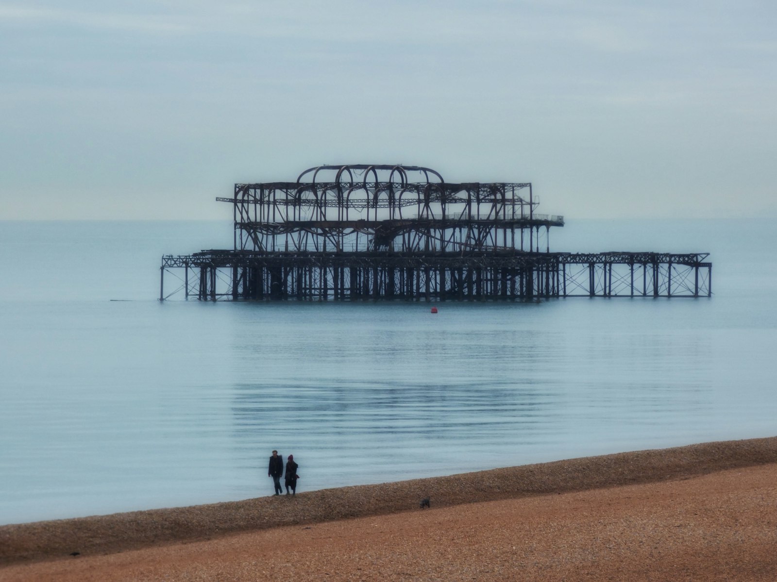 Brighton pebble beach with the old West Pier out in the calm sea, a couple walking by the shore